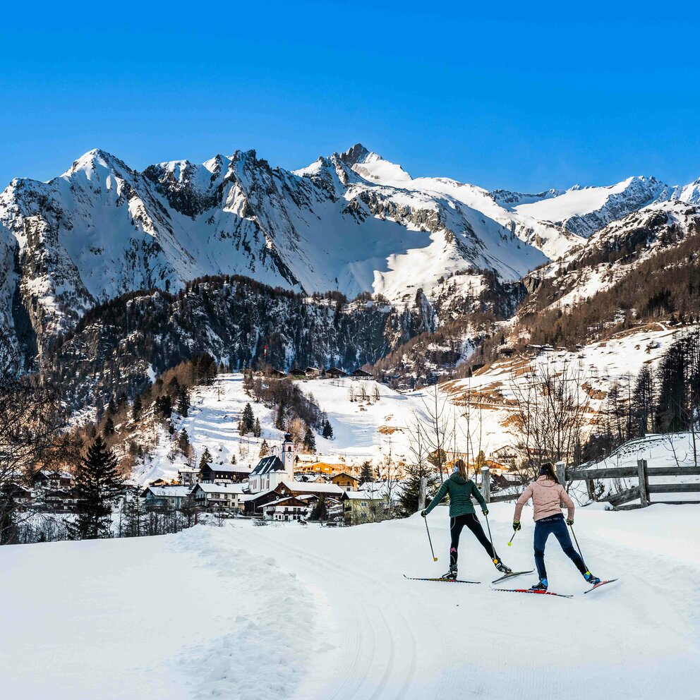 Zwei Frauen am Langlaufen mit Panoramablick auf die Gemeinde Prägraten am Großvenediger. Das verschneite Dorf wird von der Abendsonne angeleuchtet.