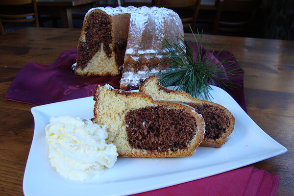 Zwei Stück Marmor-Gugelhupf auf einem Teller mit Schlag und dem ganzen Gugelhupf im Hintergrund.