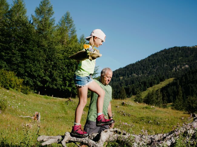 Ein kleines Mädchen balanciert mit Hilfe ihres Großvaters über einen dicken abgebrochenen Ast eines Baumes.