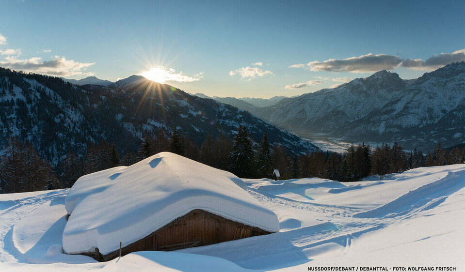 Die unberührte Schneelandschaft im Debanttal, eine Hütte liegt unter einer dicken Schneeschicht und die Sonne glänzt hinter dem Horizont hervor. 