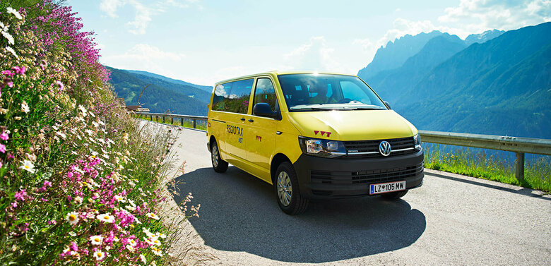 Gratisbus Ein gelber VW Bus fährt auf einer Straße, rechts sieht man eine Böschung mit blühenden Sommerblumen