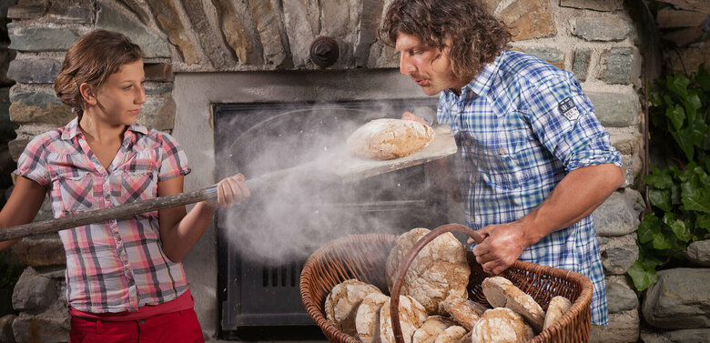 Urlaub am Bauernhof Ein Mann und eine Frau beim Brot backen. Die Frau holt ein frisch gebackenes Brot aus dem Ofen. Der Mann hält einen Korb mit mehreren Laib Brot.