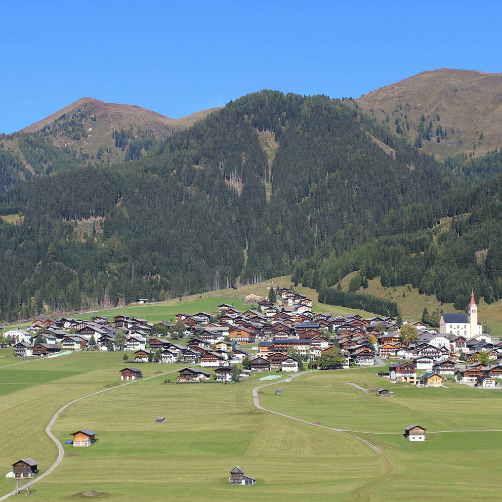 Blick von Süden auf den großen Schwemmkegel im Sommer, der den Obertilliacher Ortskern auf sich trägt. Hinter dem Dorf erheben sich die bewaldeten Hänge bis zur Baumgrenze, wo sich dann die Bergrücken der Gailtaler Alpen in die Landschaft strecken.