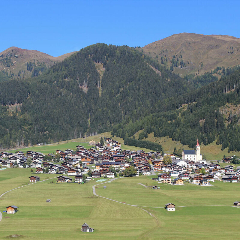 Blick von Süden auf den großen Schwemmkegel im Sommer, der den Obertilliacher Ortskern auf sich trägt. Hinter dem Dorf erheben sich die bewaldeten Hänge bis zur Baumgrenze, wo sich dann die Bergrücken der Gailtaler Alpen in die Landschaft strecken.