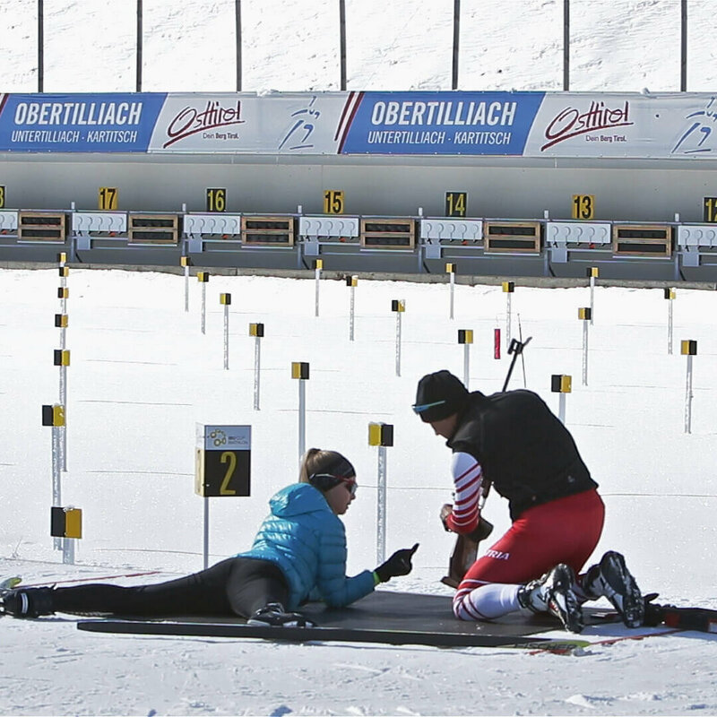 Biathlonschießen - Obertilliach Eine Frau versucht sich am Schießstand im Biathlonzentrum Obertilliach am Schießen. Dabei wird sie von einer rechts neben ihr knieenden Person unterstützt.