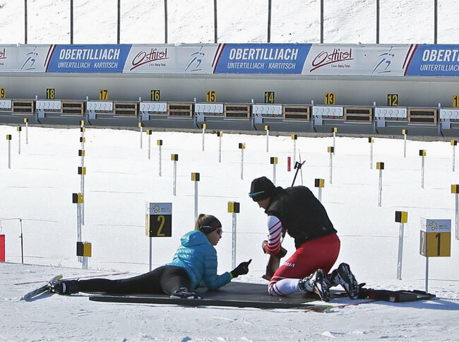 Biathlonschießen - Obertilliach Eine Frau versucht sich am Schießstand im Biathlonzentrum Obertilliach am Schießen. Dabei wird sie von einer rechts neben ihr knieenden Person unterstützt.