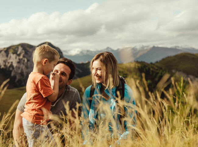 Zu sehen ist ein Elternpaar mit ihrem kleinen Sohn, die im hohen Gras entlang der Kindermeile am Golzentipp den Moment zu dritt genießen.