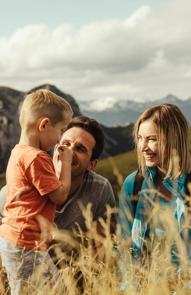 kindermeile-obertilliach Zu sehen ist ein Elternpaar mit ihrem kleinen Sohn, die im hohen Gras entlang der Kindermeile am Golzentipp den Moment zu dritt genießen.