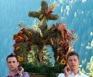 Zwei Männer tragen ein Gesteck aus Heu, Blumen und Obst zum Erntedank.