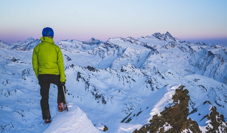Ein Skitourengeher mit grüner Jacke und blauem Helm steht auf dem Gipfel der Weißspitze und blickt in den Abendhimmel über den schneebedeckten Berggipfeln.