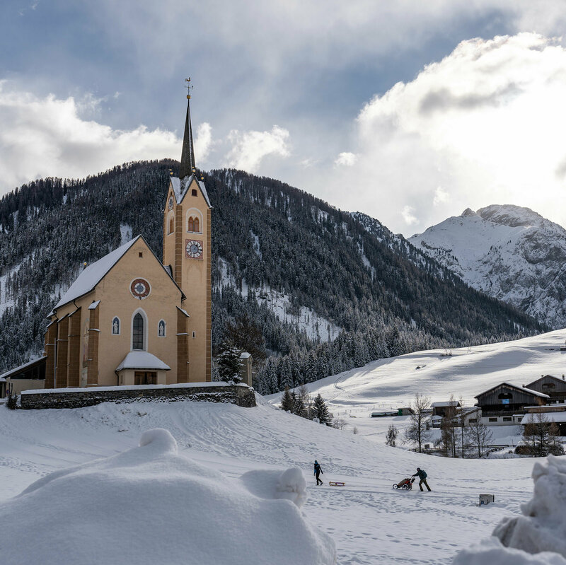 Kartitscher Pfarrkirche im Winter umgeben von schneebedeckten Feldern und den Karnischen Alpen im Hintergrund