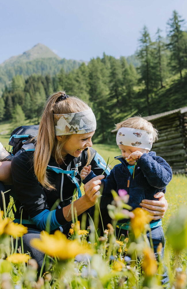 Eine kleine Familie ist im Zuge einer Familienwanderung auf einer Wiese im Kristeinertal. Die Frau und das Kind tragen Stirnbänder und der Mann hat einen roten Trage-rucksack auf dem Rücken. Sie sitzen im grünen Gras.