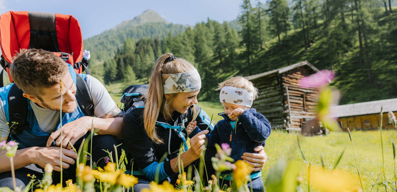 Eine kleine Familie ist im Zuge einer Familienwanderung auf einer Wiese im Kristeinertal. Die Frau und das Kind tragen Stirnbänder und der Mann hat einen roten Trage-rucksack auf dem Rücken. Sie sitzen im grünen Gras.
