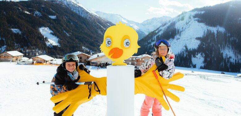 Zwei Kinder stehen im familienfreundlichen Skigebiet Golzentipp in Obertilliach hinter einem gelben Kurvenzeiger in Entendesign mit winterlichem Bergpanorama im Hintergrund.