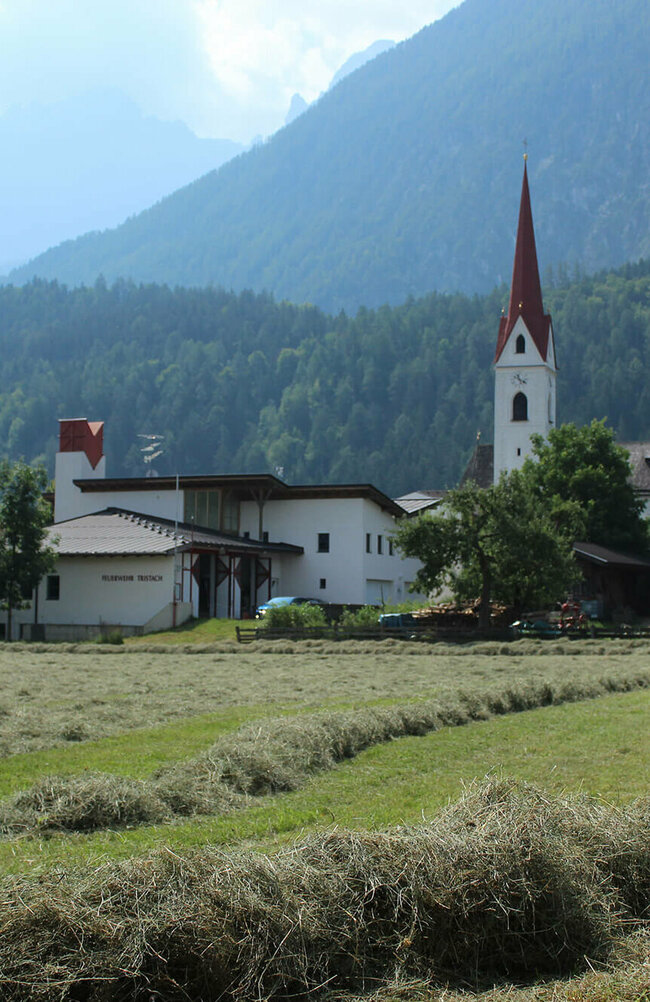Die Tristacher Kirche, das Feuerwehr Gebäude und ein abgemähtes Feld mit liegendem Heu.