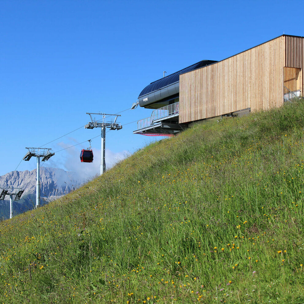 Blumenreiche, sattgrüne Bergwiese mit Seilbahn-Bergstation in senkrecht verlaufender, leicht verwitterter Holzverkleidung. Die letzten drei Liftstützen vor der Bergstation und eine rote Seilbahngondel sowie der blaue Himmel und im Hintergrund eine halbrunde, markante Felsburg bilden einen schönen Kontrast dazu.