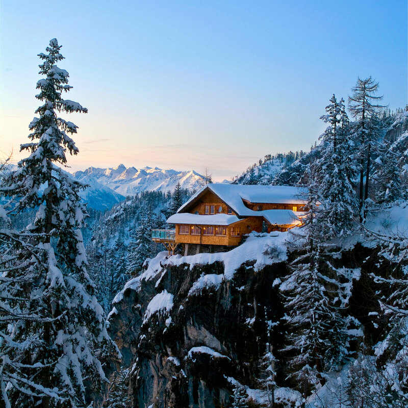 Die Dolomitenhütte steht märchenhaft in der winterlichen Abenddämmerung auf einem Plateau vor einem steilen Abhang.