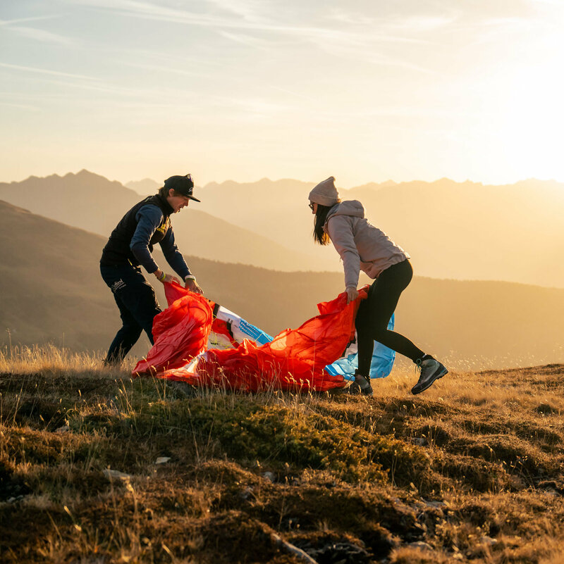 Ein Mann und eine Frau stehen auf einer Bergwiese in Osttirol in der Morgensonne und falten einen roten Gleitschirm zusammen.