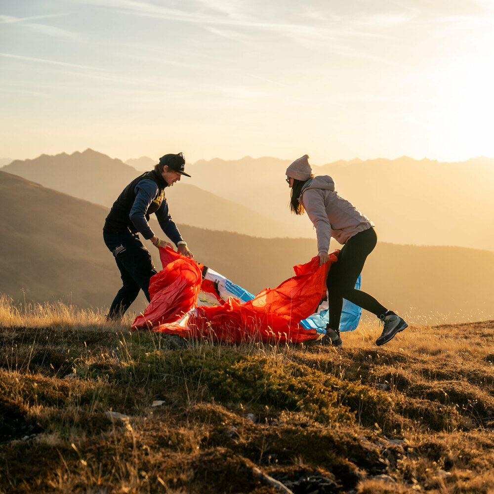 Ein Mann und eine Frau stehen auf einer Bergwiese in Osttirol in der Morgensonne und falten einen roten Gleitschirm zusammen.