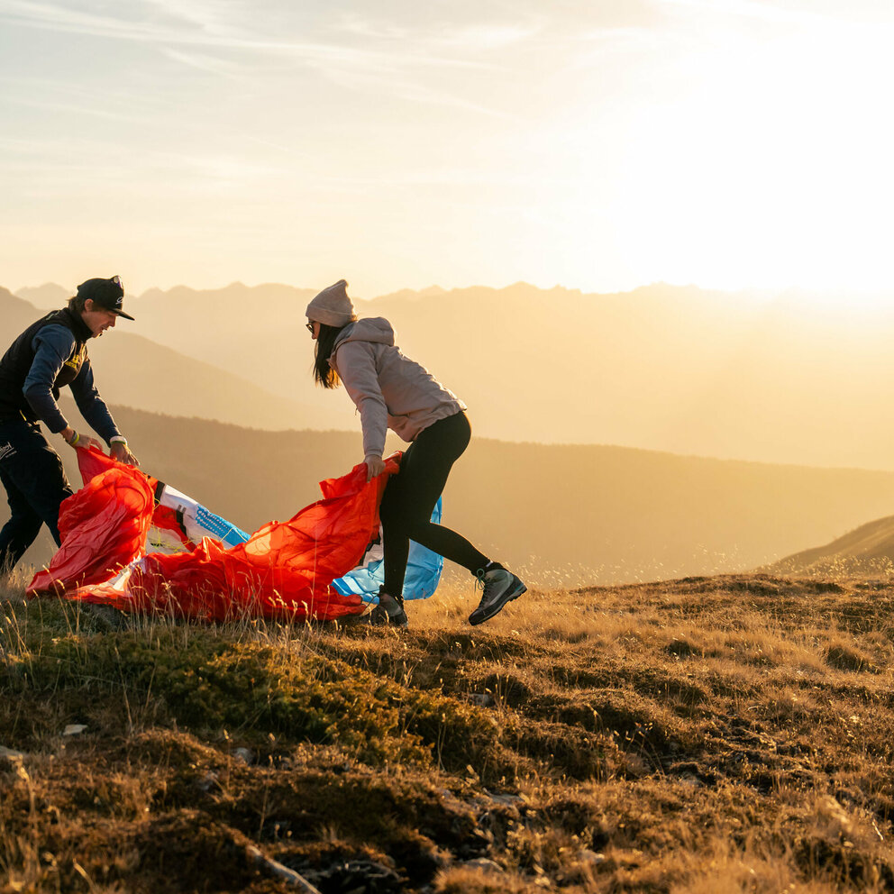 Ein Mann und eine Frau stehen auf einer Bergwiese in Osttirol in der Morgensonne und falten einen roten Gleitschirm zusammen.