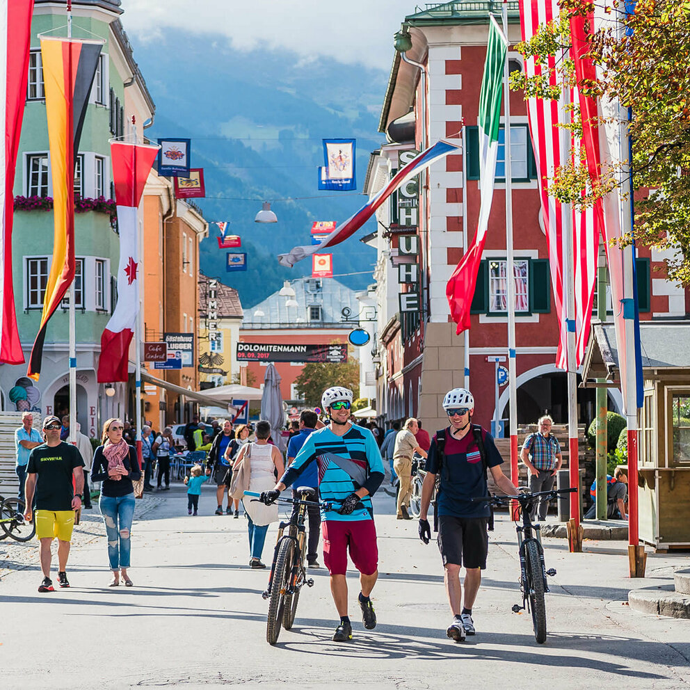 Zwei Mountainbiker schieben ihre Räder durch die mit Fahnen dekorierte Innenstadt von Lienz.