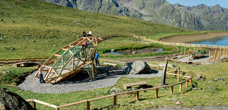 Zu sehen ist eine Familie mit Kindern die auf einem Spielgerät turnen, welches einem überdimensional-großen Frosch ähnlich sieht. Dieses Spielgerät befindet sich beim Wassermythos Ochsenlacke im Skizentrum St. Jakob i. D.. Der sichtbare Teil des Ufers, die Umgebung und das dahinterliegende Bergpanorama erstrahlen in einem sonnig-warmen Licht.