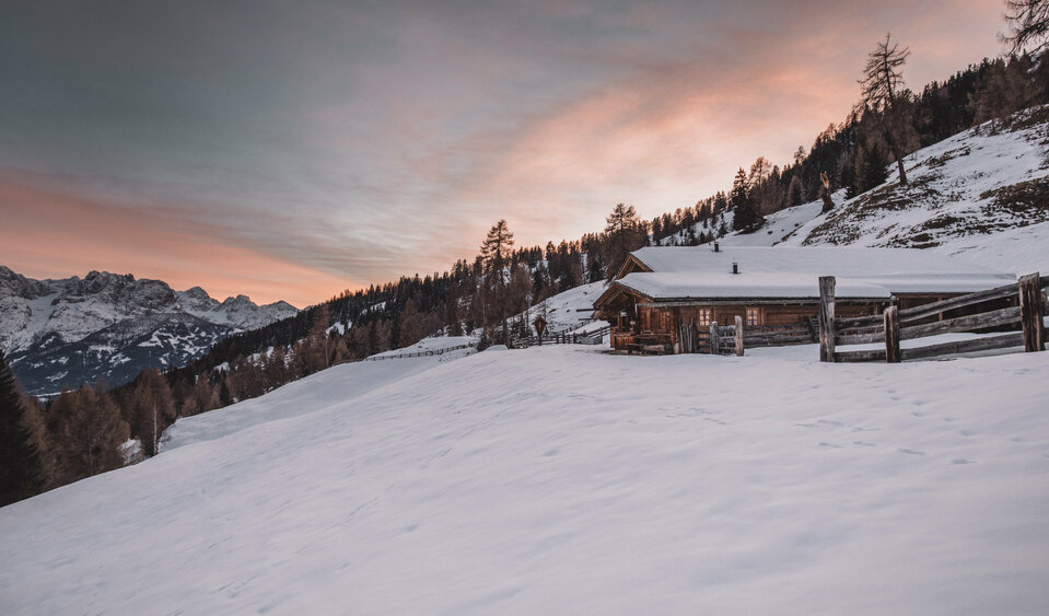 Verschneite Grissmannalm im Winter mit Wald im Hintergrund und einem rötlichen Abendhimmel.