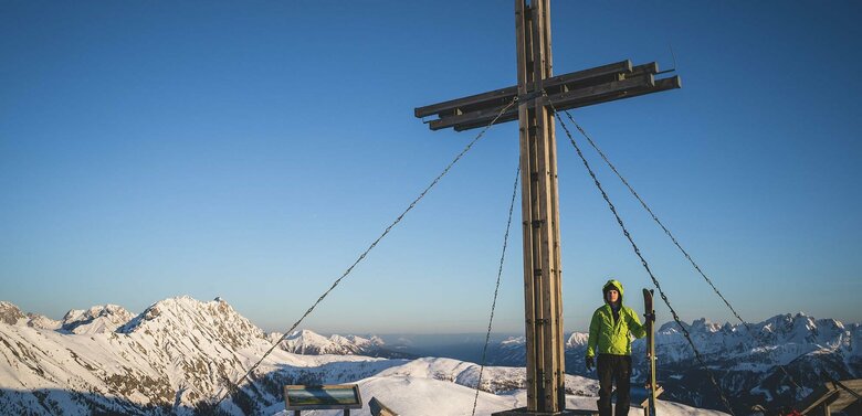 Skitourengeher beim Gipfelkreuz des Golzentipps in Obertilliach mit Blick auf die umliegende Bergwelt.