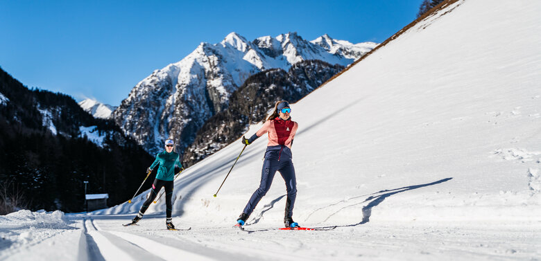 Aufnahme von unten von zwei skatenden Langläuferinnen über eine Loipe in Prägraten am Großvenediger mit Bergkulisse im Hintergrund.