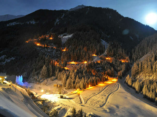 Die beleuchtete Rodelbahn Tilliachalmweg schlängelt sich in verschneiten Außervillgraten bei Nacht durch den Wald..