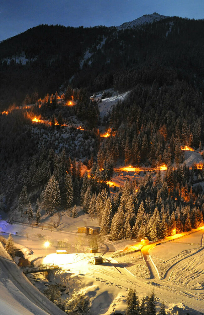 Die beleuchtete Rodelbahn Tilliachalmweg schlängelt sich in verschneiten Außervillgraten bei Nacht durch den Wald..
