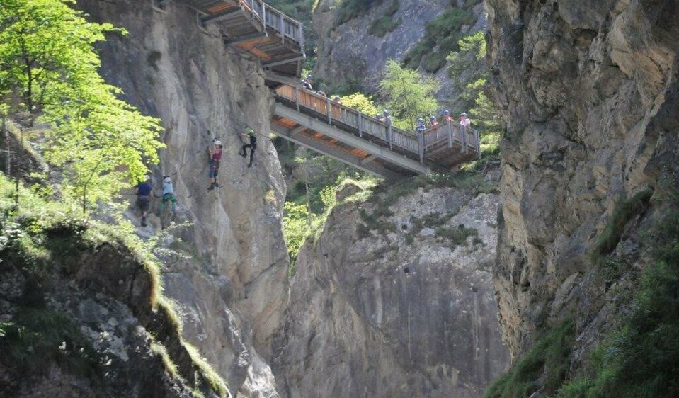 Vier Personen wagen sich mutig auf den Klettersteig der Galitzenklamm, unter ihnen tost der Bach. Von oben beobachten neugierige Besucher das Geschehen vom Wasserschaupfad aus.
