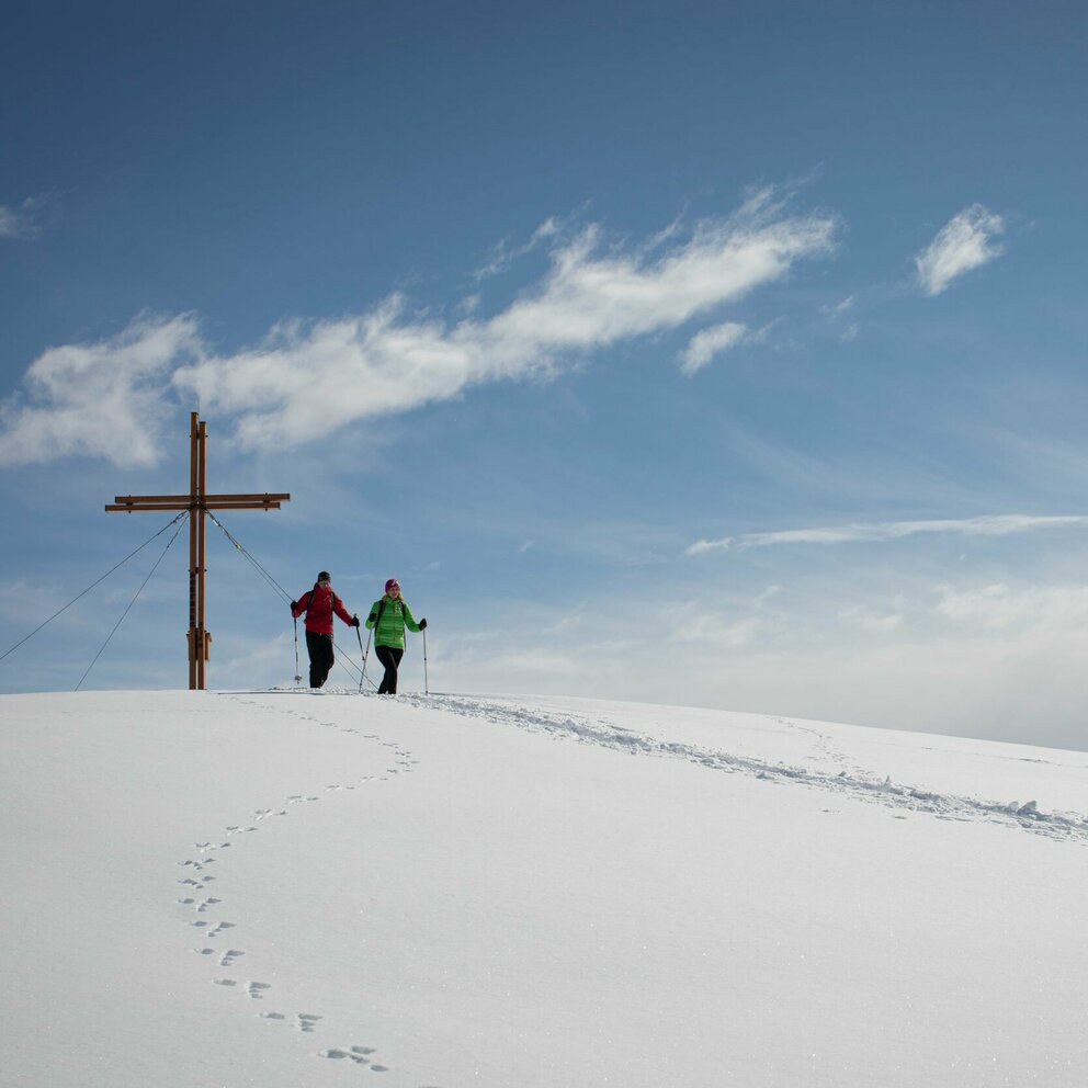 Zwei Personen stehen beim Gipfelkreuz des Dorfbergs umgeben von einer bezaubernden Winterlandschaft. Im Schnee sind außerdem Tierspuren zu erkennen.
