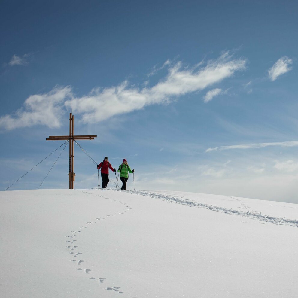Zwei Personen stehen beim Gipfelkreuz des Dorfbergs umgeben von einer bezaubernden Winterlandschaft. Im Schnee sind außerdem Tierspuren zu erkennen.