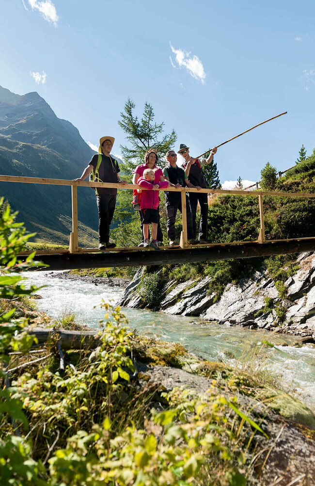 Rangertour Ein Ranger zeigt einer Familie den Nationalpark Hohe Tauern bei einer geführten Rangertour. Die Gruppe steht auf einer hölzernen Brücke über einem Fluss und im Hintergrund ragt eine Bergkette empor.