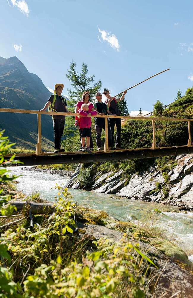 Ein Ranger zeigt einer Familie den Nationalpark Hohe Tauern bei einer geführten Rangertour. Die Gruppe steht auf einer hölzernen Brücke über einem Fluss und im Hintergrund ragt eine Bergkette empor.
