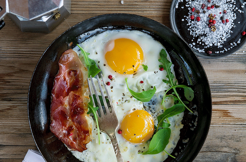 Osttirol Frühstück Zwei gebratenen Spiegeleier mit Schnittlauch und einer gebratenen Speckscheibe in einer Eisenpfanne mit frischen Tomaten auf der Rispe davor auf einem weißen Tuch schön angerichtet.