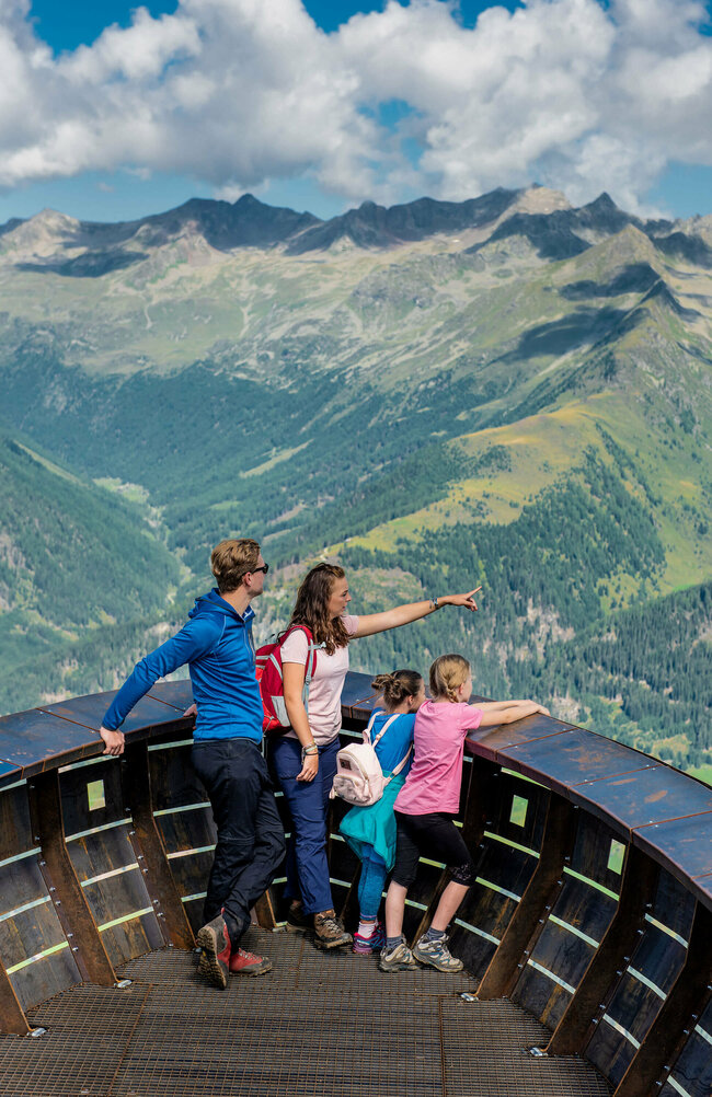 Eine vierköpfige Familie genießt den Ausblick bei der Aussichtsplattform des Wassermythos Ochsenlacke im Skizentrum St. Jakob i. D.. Ihr Blick fällt auf das gegenüberliegende Bergpanorama des Defereggentals, welches trotz einiger Wolken am Himmel in der Sonne erstrahlt.