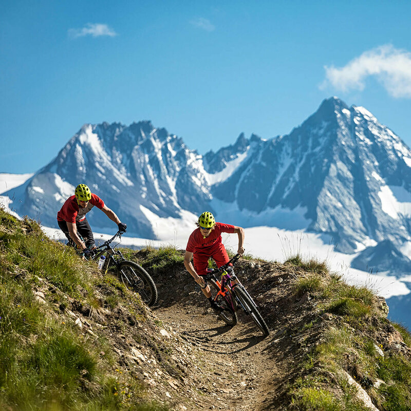 Zwei Radfahrer mit roten Shirts und gelben Helmen auf einem schmalen Trail im Bikepark Grossglockner Resort. Im Hintergrund ragt der Großglockner in den blauen Himmel.