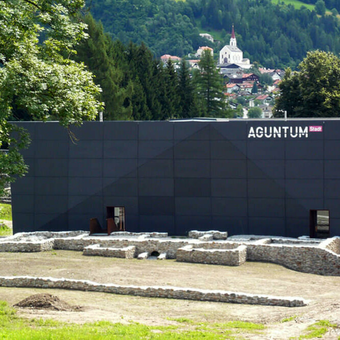 Blick auf das Museum Aguntum in Dölsach, umgeben von römischen Ausgrabungen; im Hintergrund thront auf einer Anhöhe die Kirche von Dölsach