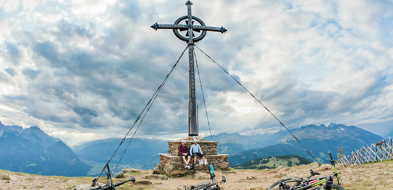 Biken Ederplan Zwei Personen sitzen am Gipfelkreuz des Ederplan. Drei Bikes liegen vor ihnen auf der Wiese.