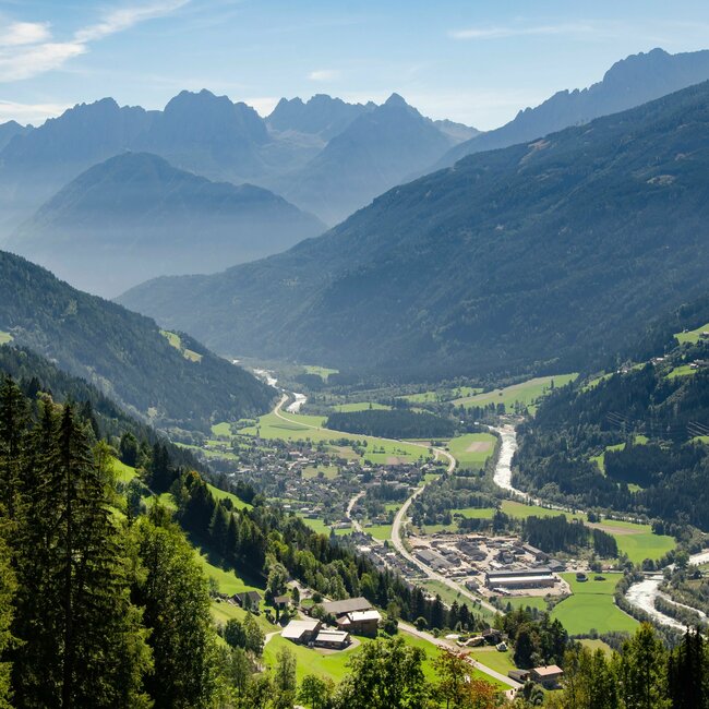 Ortsansicht Ainet im Sommer von einem höher gelegenen Aussichtspunkt mit Bergpanorama bei blauem Himmel im Hintergrund.