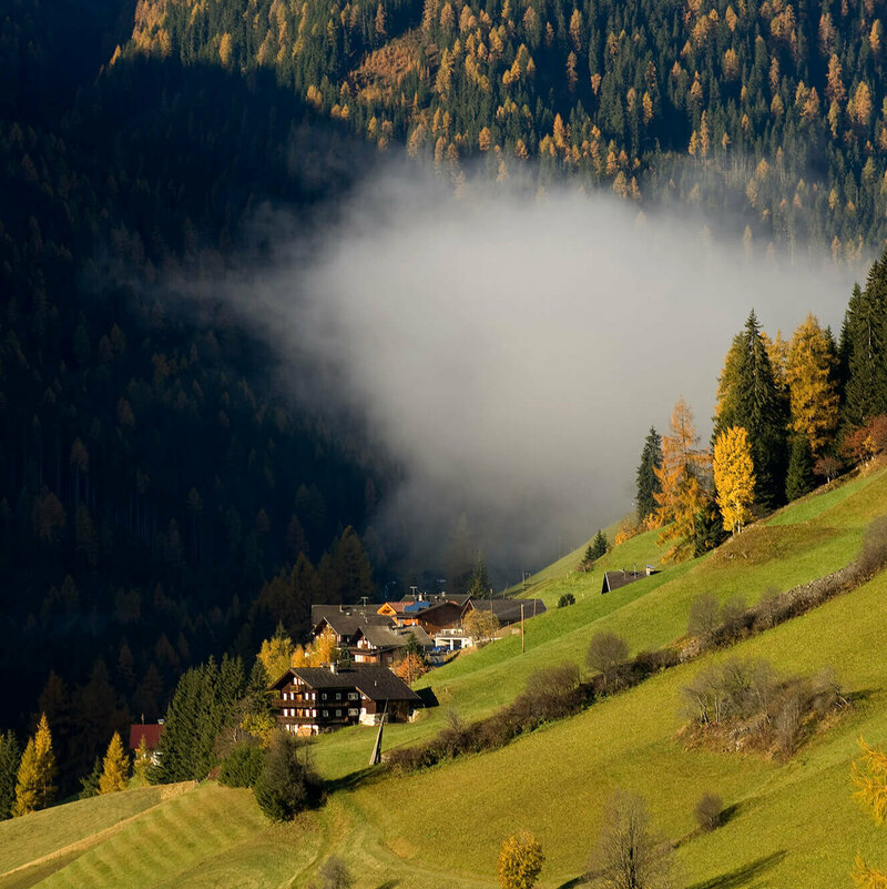 Bergbauernhöfe im herbstlichen Defereggental in Osttirol. Eine kleine Wolken hängt zwischen den Bergen und die Bäume sind bereits herbstlich gefärbt.