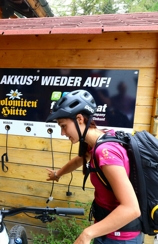 Ein Pärchen mit Fahrrädern steht vor der E-Bike Ladesäule bei der Dolomitenhütte. An einer Holzhütte hängt ein Plakat mit den Worten "Wir laden auch ihre Akkus wieder auf".