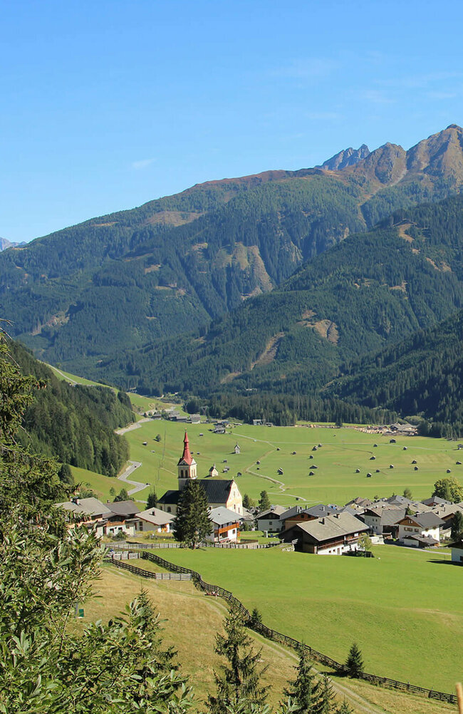 Blick auf eng verbautes Haufendorf mit ausschließlich grauer Dacheindeckung an einem wolkenlosen Herbsttag. Im Hintergrund die grünen Felder mit vielen kleinen Heuhütten und ein Gebirgszug, der das Tal mit seinen bewaldeten Hängen verschließt.