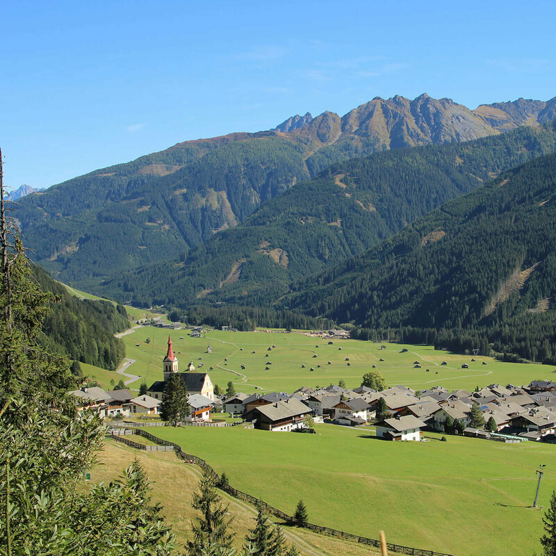 Blick auf eng verbautes Haufendorf mit ausschließlich grauer Dacheindeckung an einem wolkenlosen Herbsttag. Im Hintergrund die grünen Felder mit vielen kleinen Heuhütten und ein Gebirgszug, der das Tal mit seinen bewaldeten Hängen verschließt.