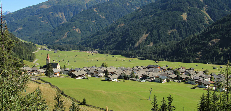 Obertilliach Blick auf eng verbautes Haufendorf mit ausschließlich grauer Dacheindeckung an einem wolkenlosen Herbsttag. Im Hintergrund die grünen Felder mit vielen kleinen Heuhütten und ein Gebirgszug, der das Tal mit seinen bewaldeten Hängen verschließt.