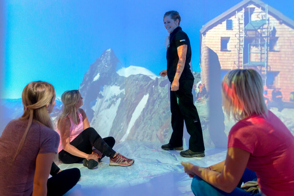 Drei Frauen sitzen aufmerksam am Boden und lauschen der Nationalpark-Rangerin, die mit einem Finger auf den majestätischen Großglockner weist.