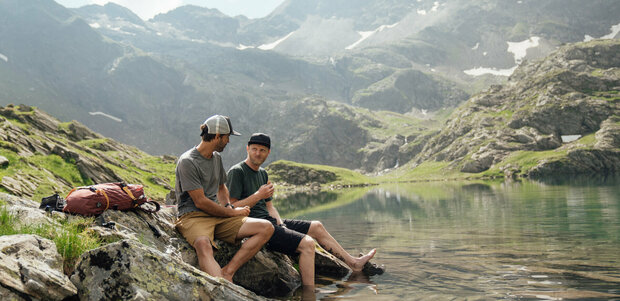 Zwei Männer sitzen am Geigensee und genießen das frische und kristallklare Wasser. Dabei essen sie ihre Jause und unterhalten sich.