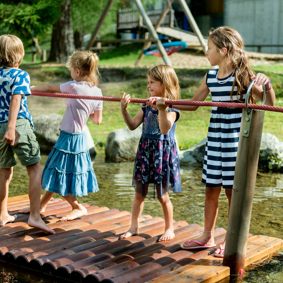 Vier Kinder stehen auf einer Plattform im See auf dem Wassernaturspielplatz in Sankt Jakob und halten sich an einem roten Seil fest. 
